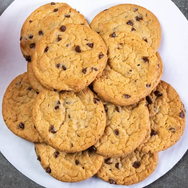 A close-up, overhead view of a pile of golden-brown chocolate chip cookies on a white surface.